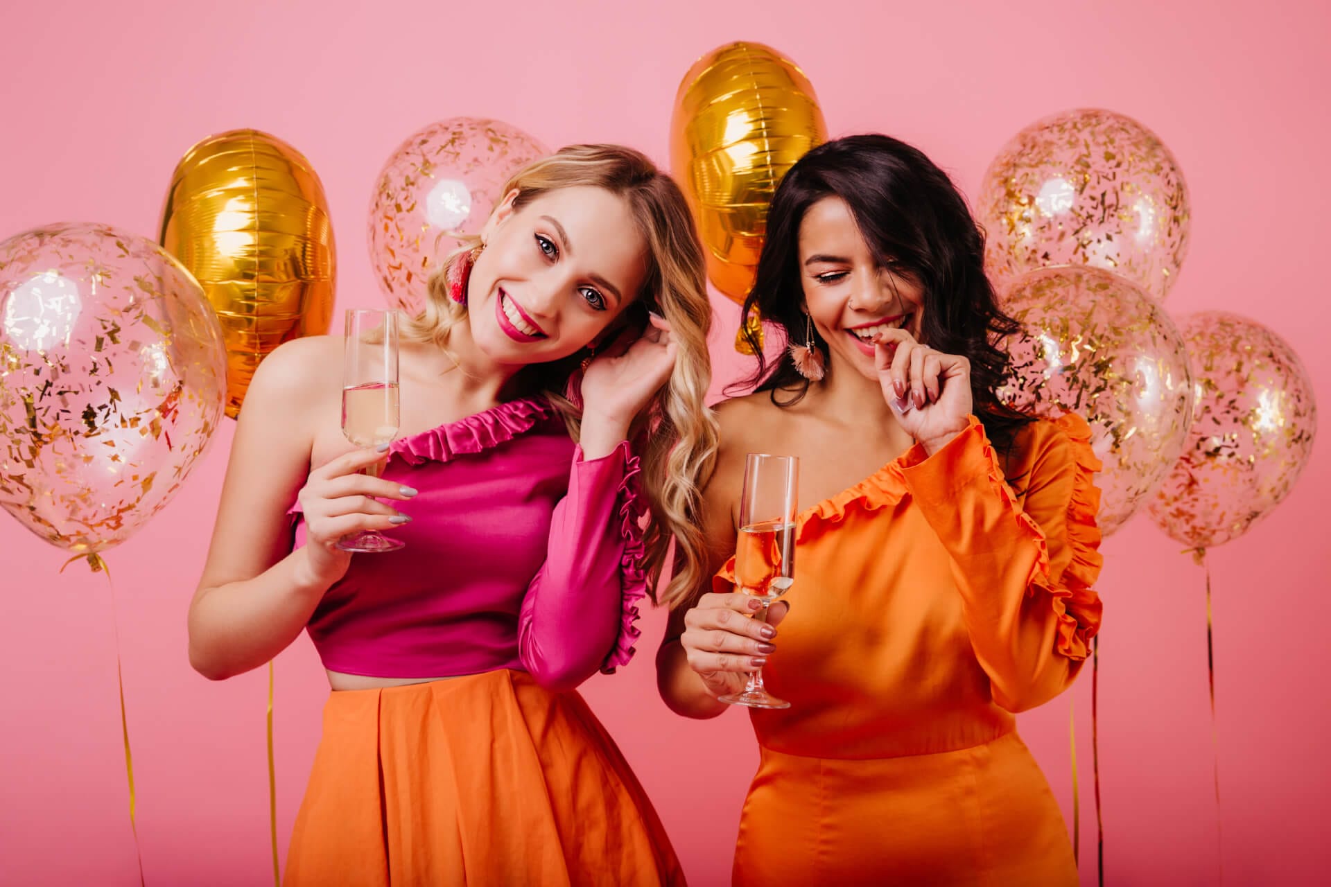 Half length portrait of two young ladies drinking champagne. Indoor photo of girls with balloons isolated on pink background