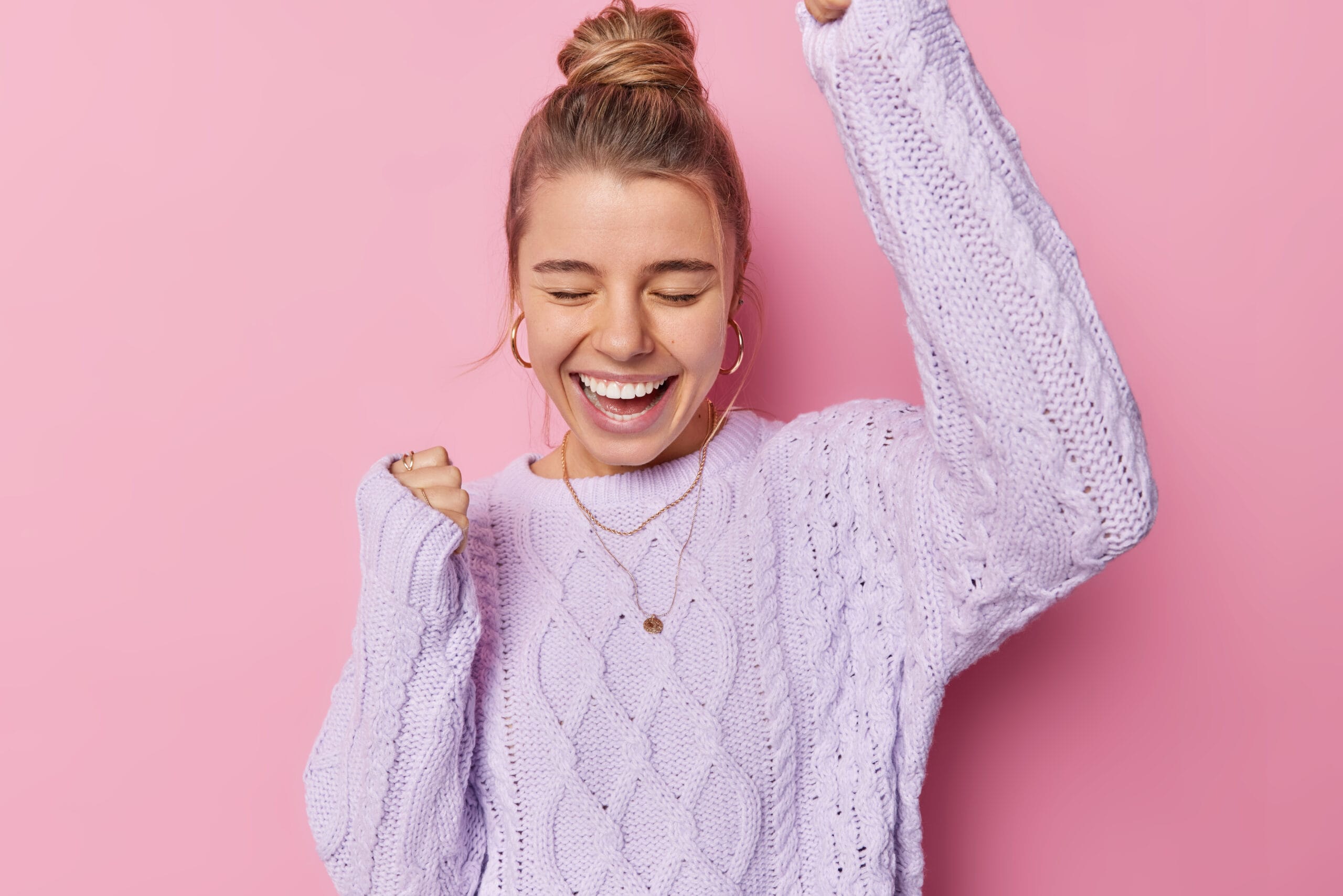 Indoor shot of optimistic young woman with bun hairstye makes yes gesture laughs positively celebrates achievements wears knitted purple jumper isolated over pink background. Finally success